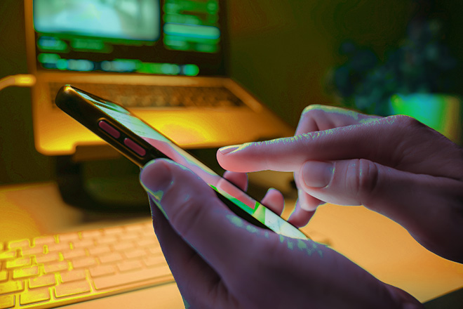 Image of hand holding a smartphone with a laptop in the background.